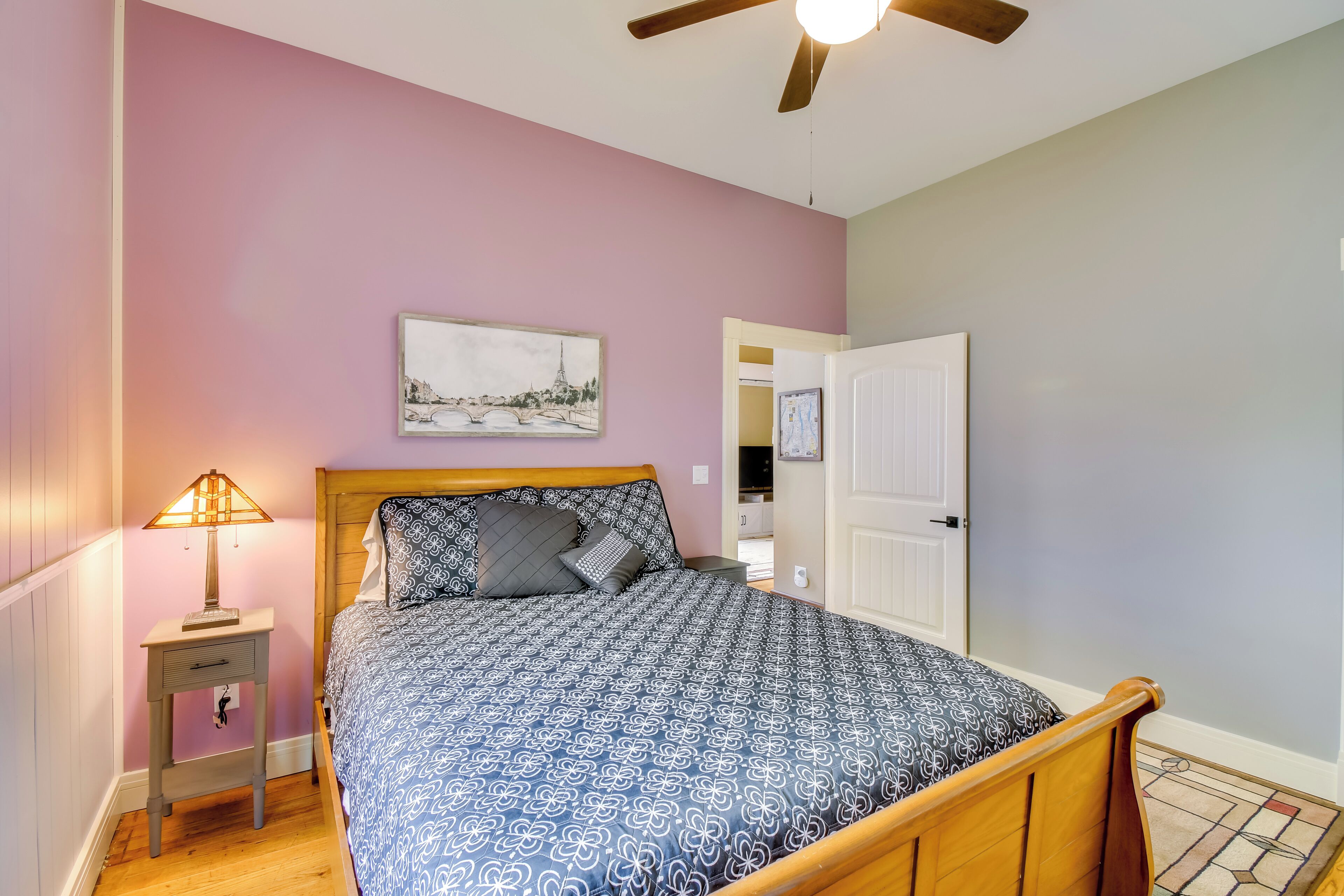 A bedroom with a bed, a nightstand, a lamp, a ceiling fan, and a door leading to another room.