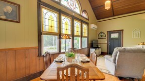 A dining area with a wooden table set for four, surrounded by wooden chairs, large stained glass windows, and a grey sofa. - Restored Ovid Church Near Cayuga Lake Wine Trail (Ovid)