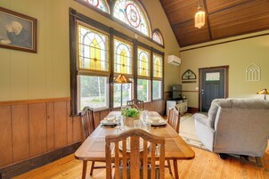 Un comedor con una mesa de madera dispuesta para cuatro personas, rodeada de sillas de madera, amplias ventanas con vidrieras y un sofá gris.