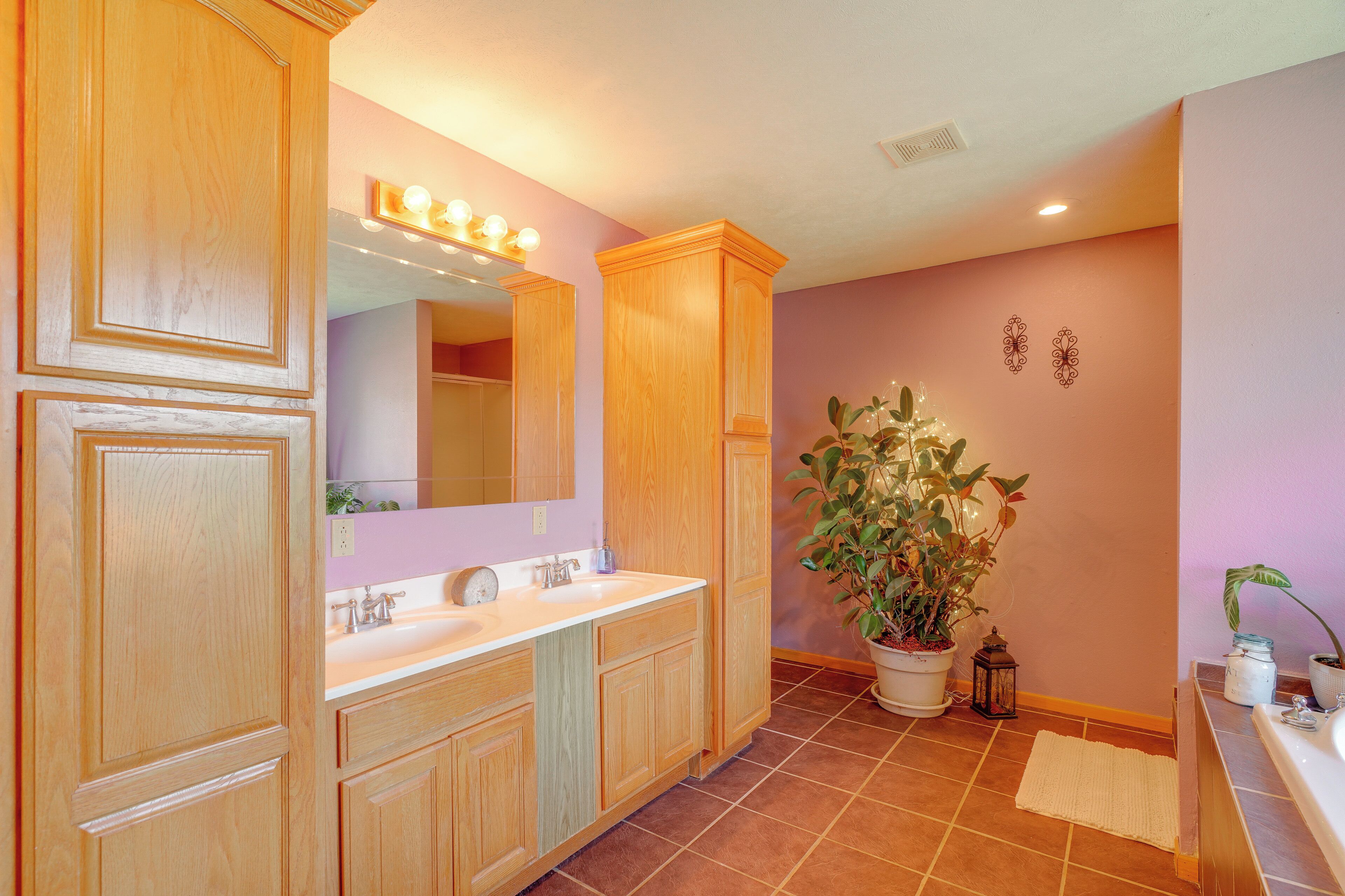 A bathroom with wooden cabinetry, a double sink vanity, a large mirror with lights, a potted plant, and a tiled floor.