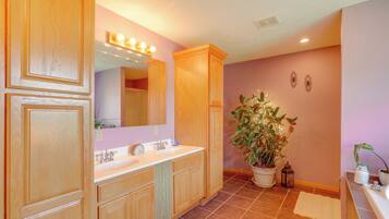 A bathroom with wooden cabinetry, a double sink vanity, a large mirror with lights, a potted plant, and a tiled floor.