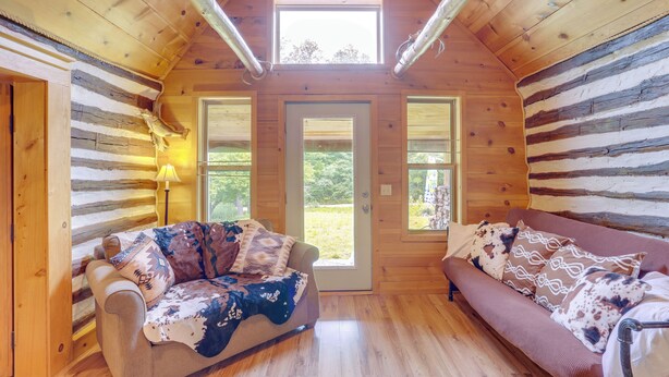 A log cabin interior with a wooden floor, two sofas with patterned cushions, a window, and a door leading outside. - Mountain-view Cabin on Farm: 3 Mi to Town & Shops (Mountain View)