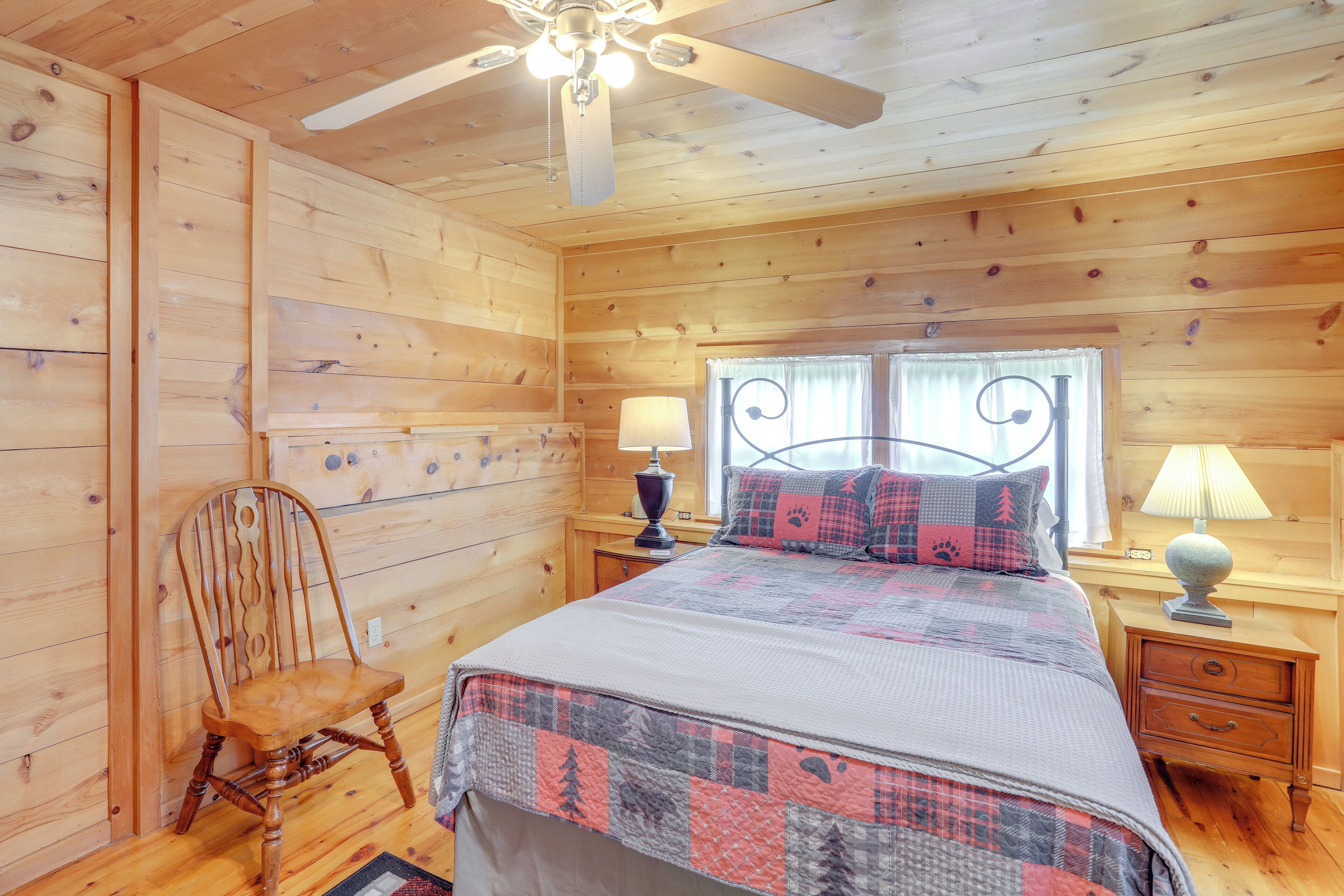 A wooden cabin bedroom with a bed, two nightstands, a chair, and a ceiling fan.