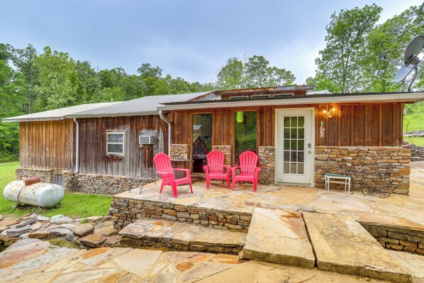 A rustic cabin with a stone foundation, a wooden exterior, and a stone patio with three Adirondack chairs.