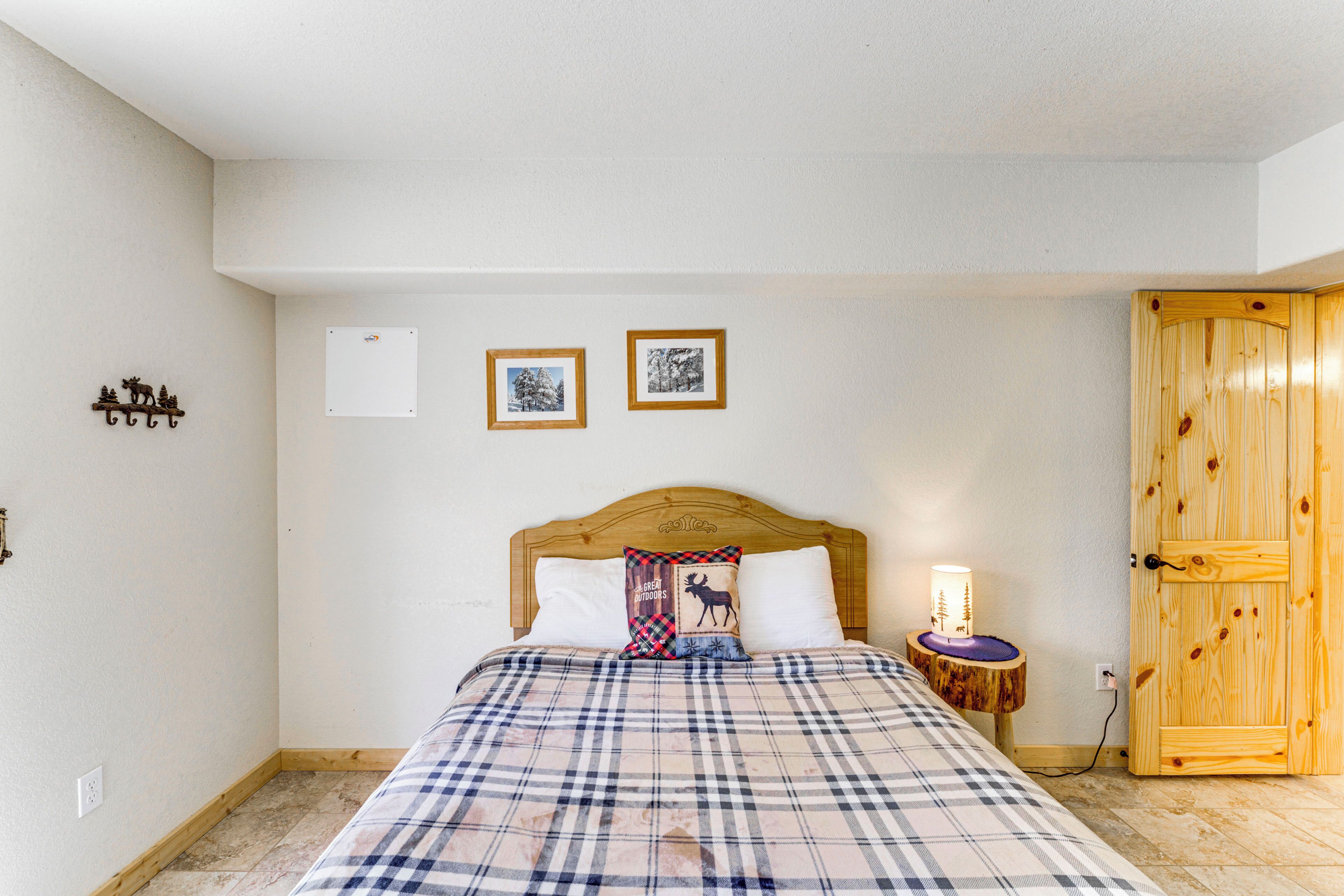Um quarto com uma cama, uma mesa de cabeceira com abajur, uma porta de madeira e duas imagens emolduradas na parede.