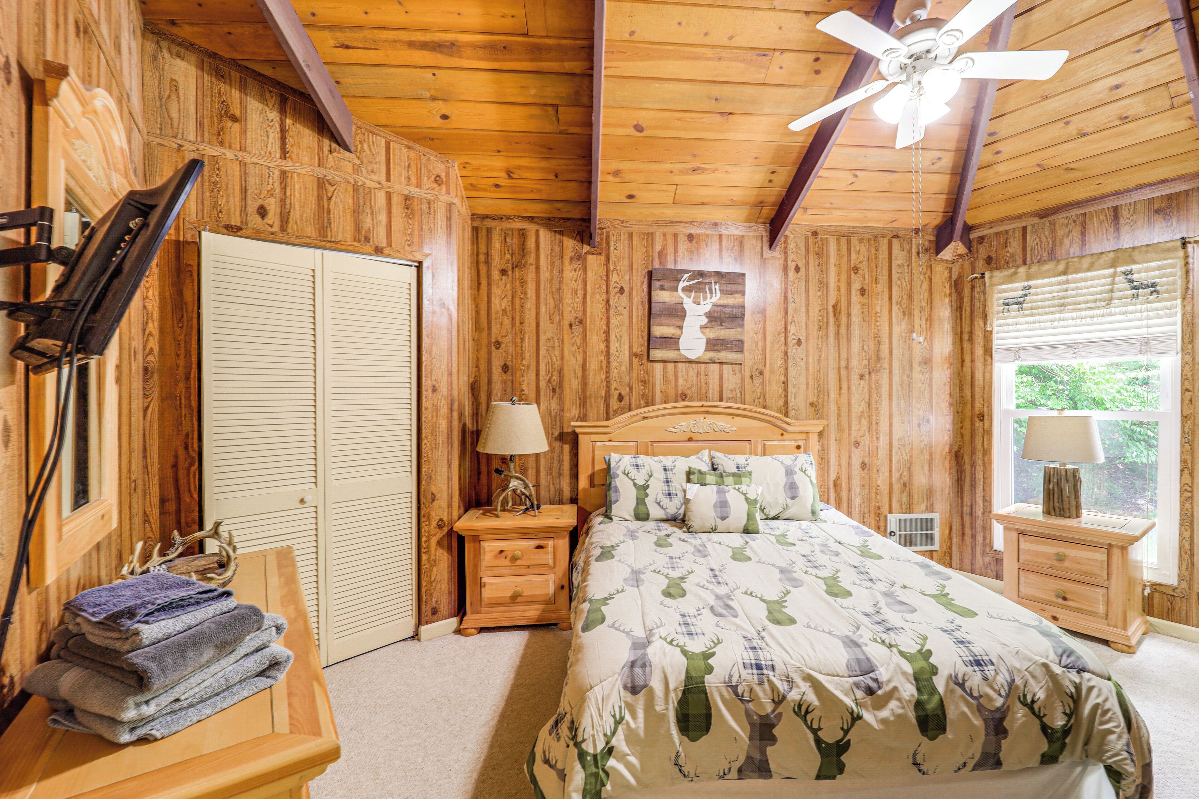 A wooden-paneled bedroom with a bed, nightstands, a ceiling fan, a window with blinds, and a wooden bench with towels.