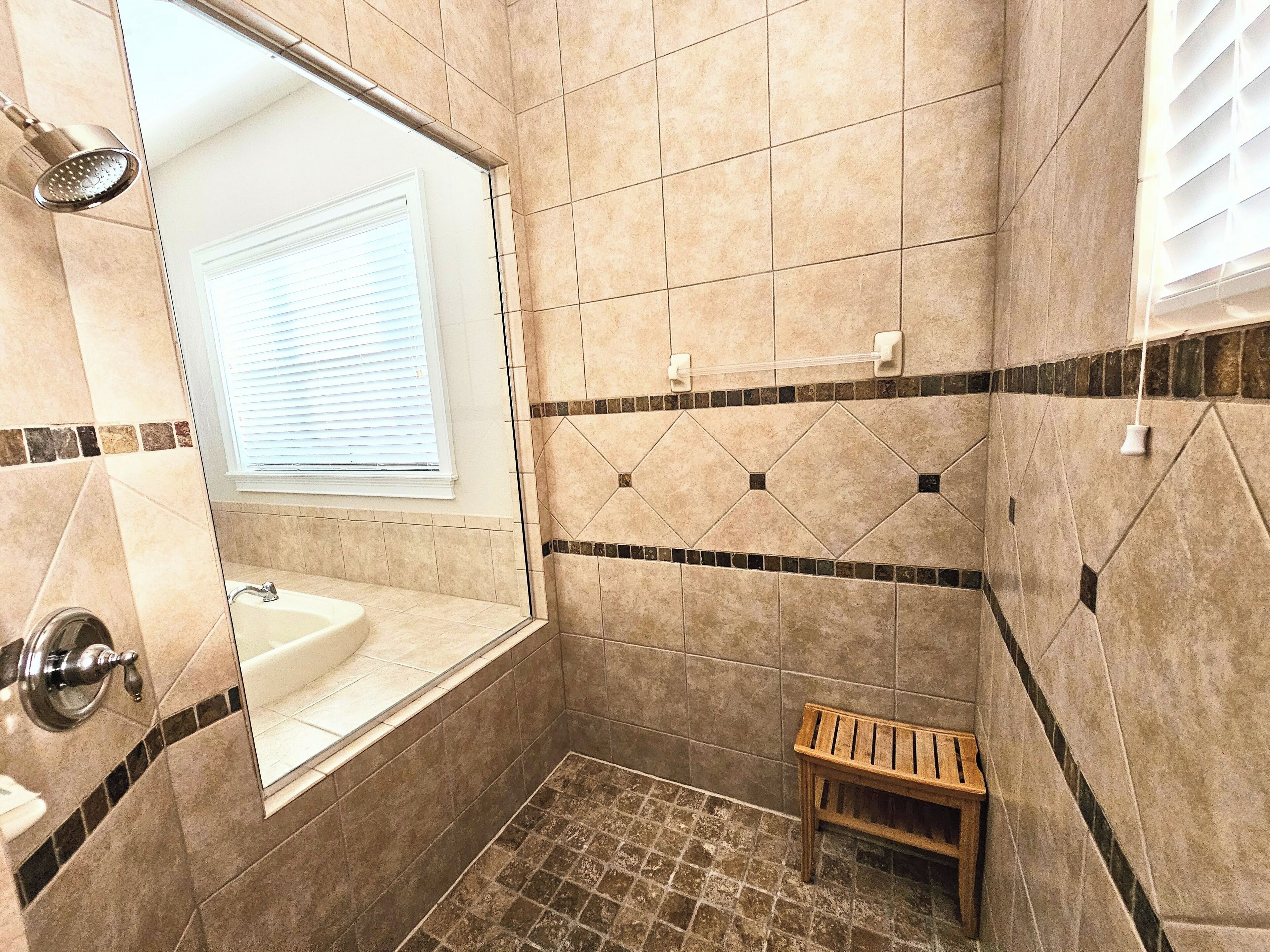 A bathroom with a bathtub, a window, a showerhead, and a small wooden bench.
