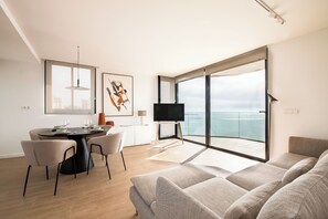 A modern living room with a large window overlooking the sea, a dining area with a round table, and a flat-screen TV on a stand. - Modern sea front Lighthouse (Alicante)