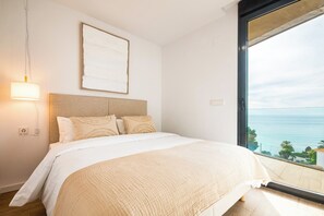 Une chambre à coucher comprenant un lit, une table de chevet, une lampe fixée au mur et une grande fenêtre donnant sur la mer.