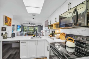 A modern kitchen with white cabinets, a black stove, and a microwave. There is a ceiling fan, a sink, and a window overlooking a balcony. - I-353 - Designer Furnished and Views & Sleeps 6 (Naples)
