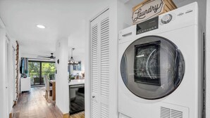 A laundry room with a large front-loading washing machine, a wooden sign above it, and a view into a dining area with a table and chairs.