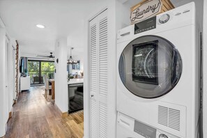 A laundry room with a large front-loading washing machine, a wooden sign above it, and a view into a dining area with a table and chairs. - I-353 - Designer Furnished and Views & Sleeps 6 (Naples)