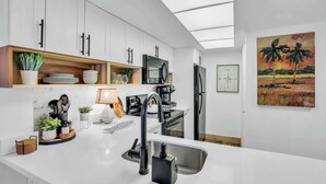 A modern kitchen with white cabinets, a black faucet, and a microwave. There is a painting of palm trees on the wall.