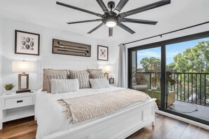 A bedroom with a large bed, a ceiling fan, a balcony with a view of trees, and a window with a curtain.