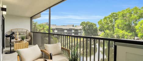 Ein Balkon mit einer Couch und einem Ventilator, der auf ein Gewässer und Grünflächen blickt.