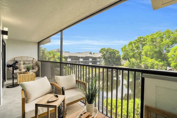 A balcony with a sofa and a fan, overlooking a body of water and greenery. - I-353 - Designer Furnished and Views & Sleeps 6 (Naples)