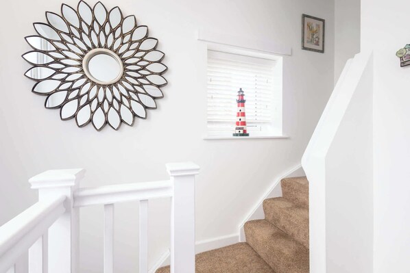 A staircase with white railings and beige carpet, a wall-mounted mirror, and a window with blinds and a small lighthouse model.