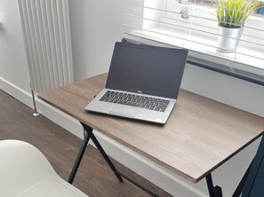A laptop on a wooden desk with a white chair in front, near a window with a plant on the sill.