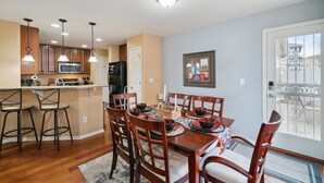 A dining area with a wooden table set for six, surrounded by chairs, in a room with a kitchen in the background. - Flyer's Nest Townhome (Westminster)