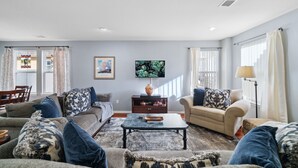 A living room with a sofa, a coffee table, and a lamp. There is a painting on the wall and a window with curtains. - Flyer's Nest Townhome (Westminster)