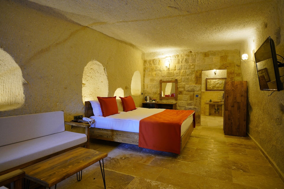 Un dormitorio con cama, un banco, una mesita de noche, un espejo, un armario de madera y un baño con lavamanos y espejo.