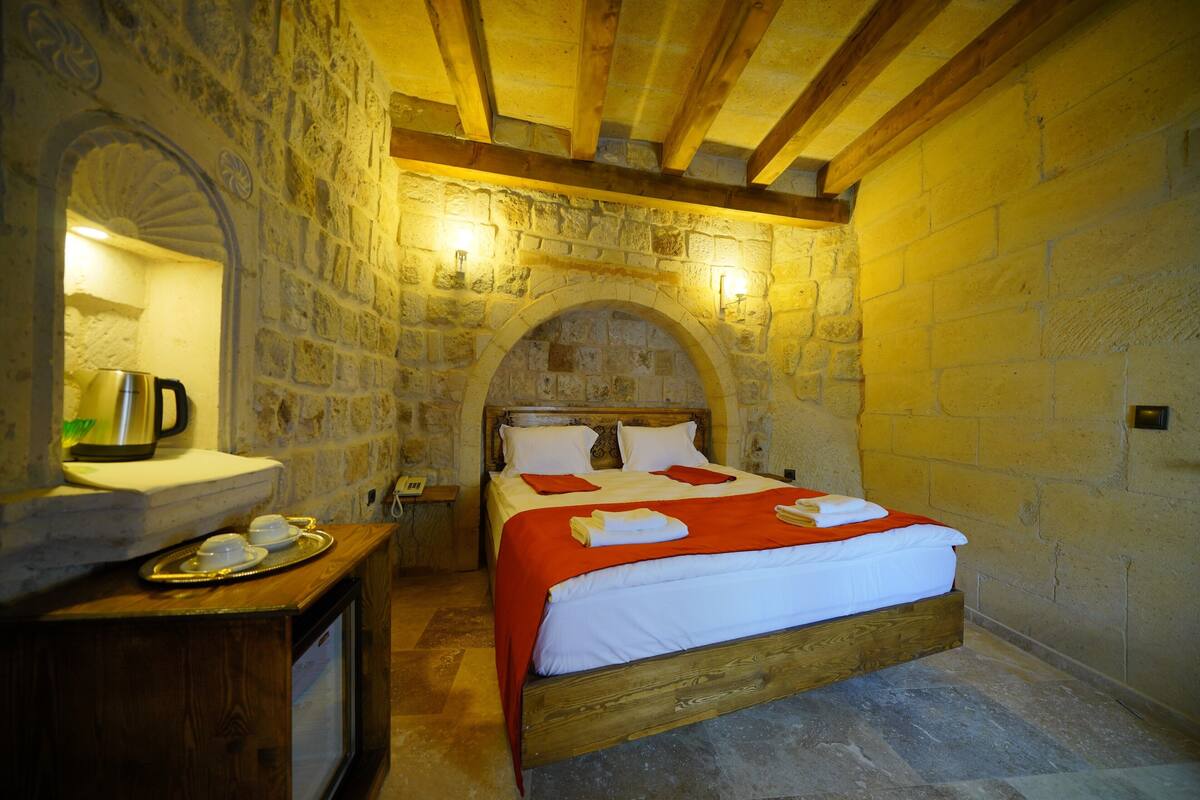 Un dormitorio de paredes de piedra con una cama de madera, un banco de piedra con una tetera y una pequeña mesa con tazas.