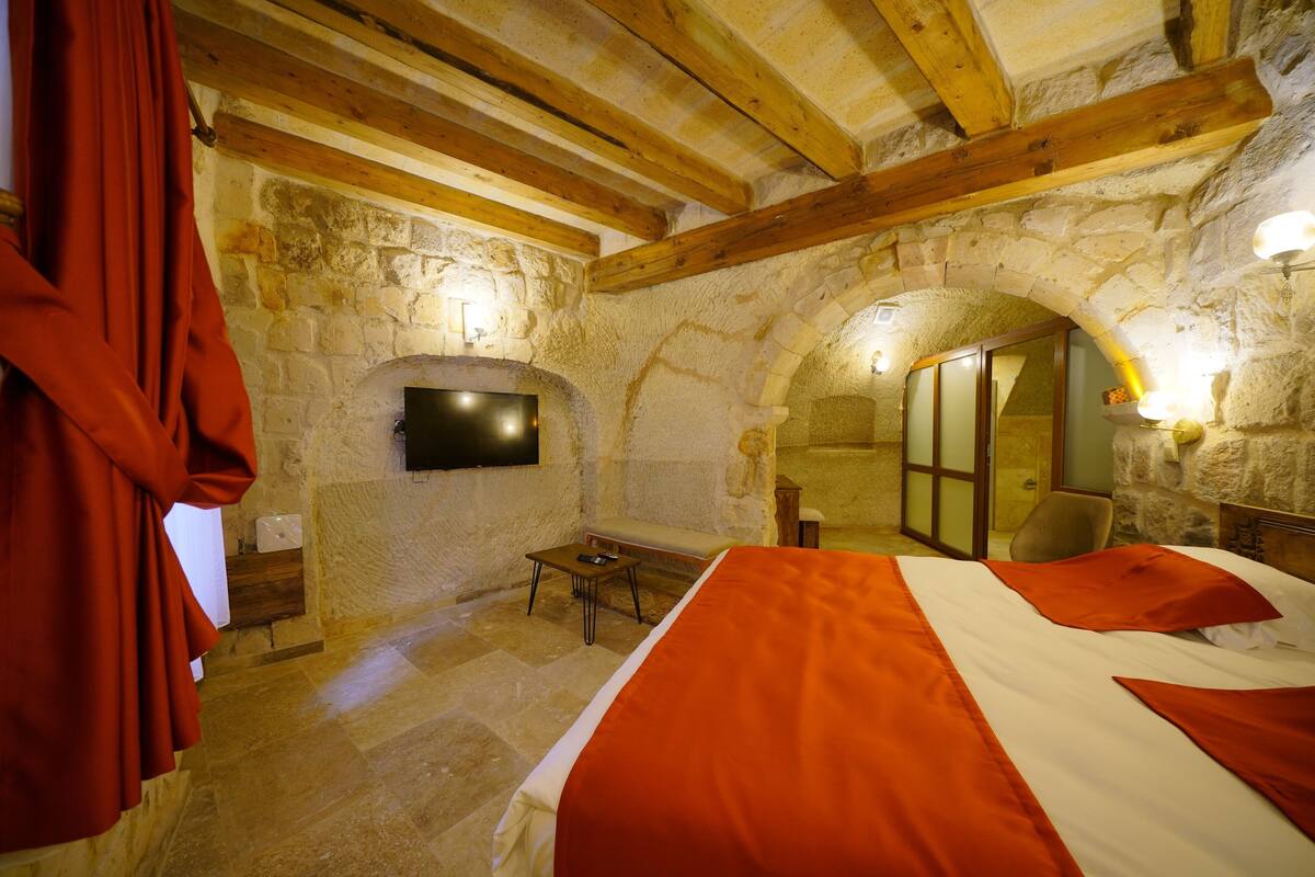 Una habitación con paredes de piedra, una cama, un televisor montado en la pared y un banco.