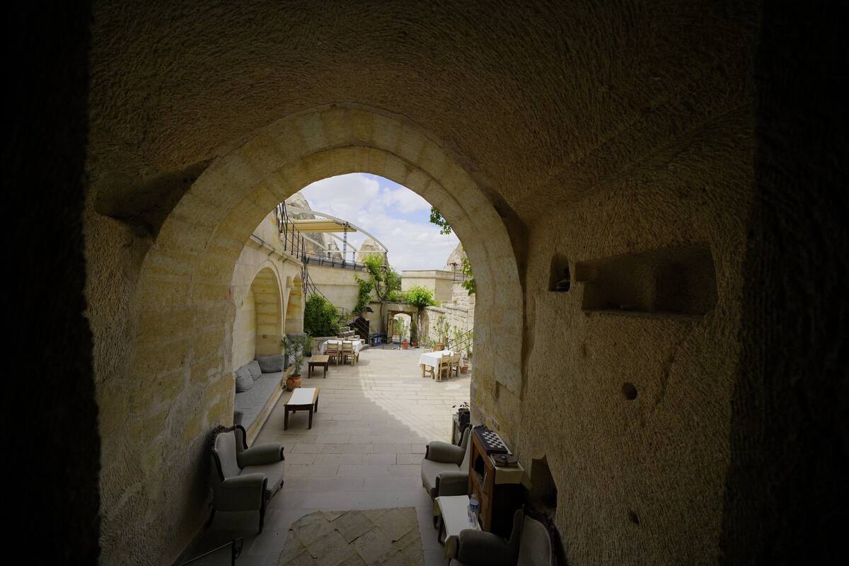 Un corredor de piedra en arco que lleva a un patio con asientos al aire libre y vistas a un edificio de arquitectura abovedada.
