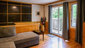 A cozy living room with a sofa, wooden walls, a large window with curtains, and a view of the outdoors.