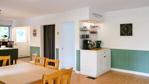 A cozy kitchen with a dining area, featuring a coffee machine, shelves with mugs, and a clock on the wall.