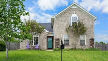 A two-story brick house with a front door, a mailbox, and a small front yard with a tree and a purple chair.