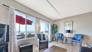 Sala de estar con televisor, una mesa de centro de madera y dos sillones azules. Hay un ventanal con un paraguas rojo y vista a la playa.