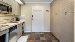 Egy kis méretű konyha, amelyben mikrohullámú sütő, mosogató és mosogatógép található. Egy fehér ajtó látható, mellette egy kabátállvány.