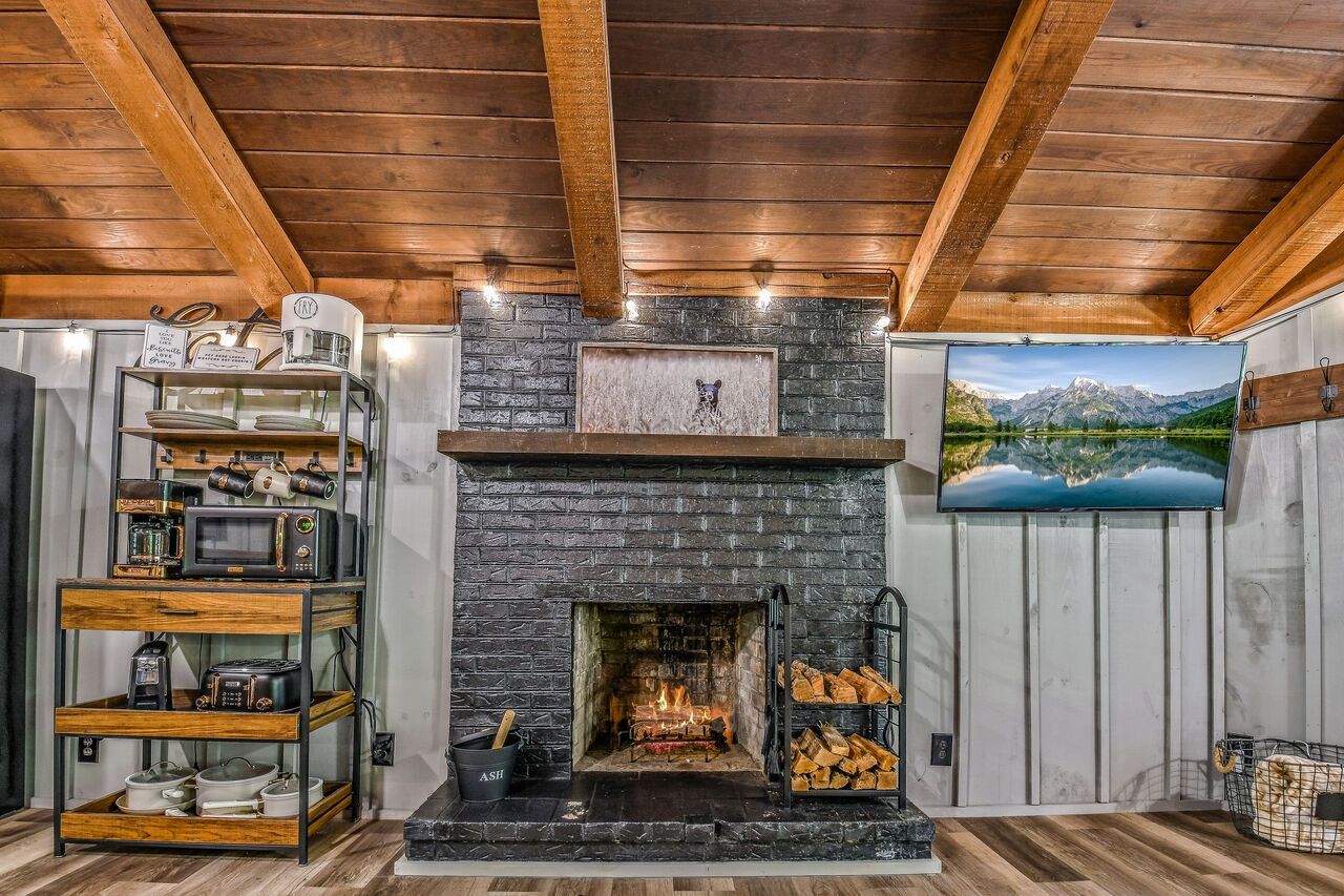 A cozy room with a fireplace, wooden beams, a TV, and a shelf with kitchen appliances.