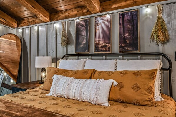A bed with multiple pillows and a headboard, a bedside lamp, and wall-mounted pictures of forests. - Light'n Bug Bungalow 1 Bedroom Cabin by RedAwning (Townsend)