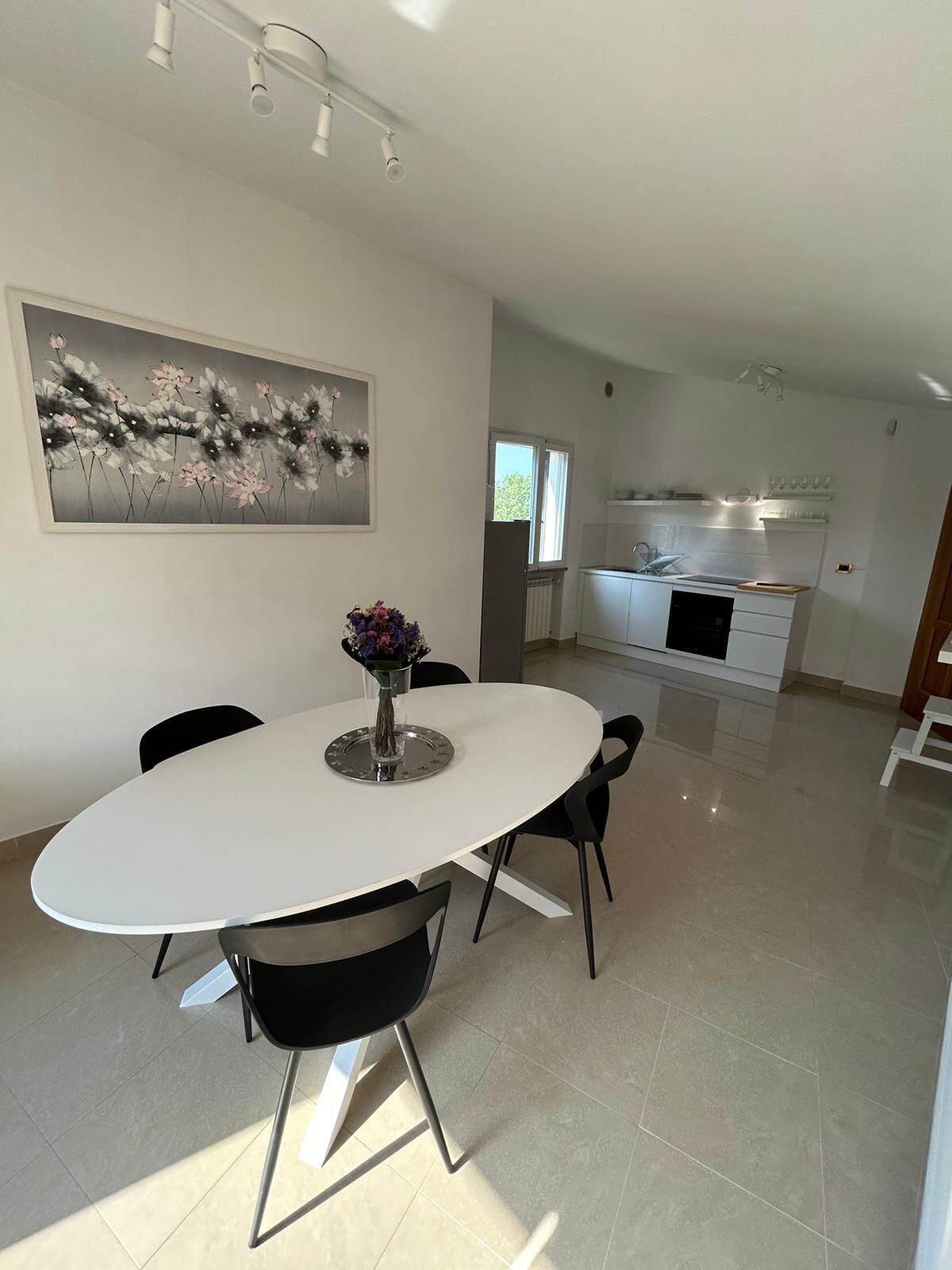 A modern dining area with a white oval table, black chairs, and a vase of flowers. A painting of flowers hangs on the wall.