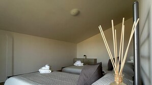 Ein modernes Hotelzimmer mit zwei Betten, einem Nachttisch mit einer Vase und Reed-Diffusern sowie einem Beistelltisch mit einer Lampe.