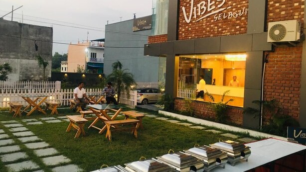 An outdoor seating area with tables and chairs, a brick building with a sign reading "Vibes Selfi", and a white fence in the background. - Hotels Vibes  (Vikasnagar)