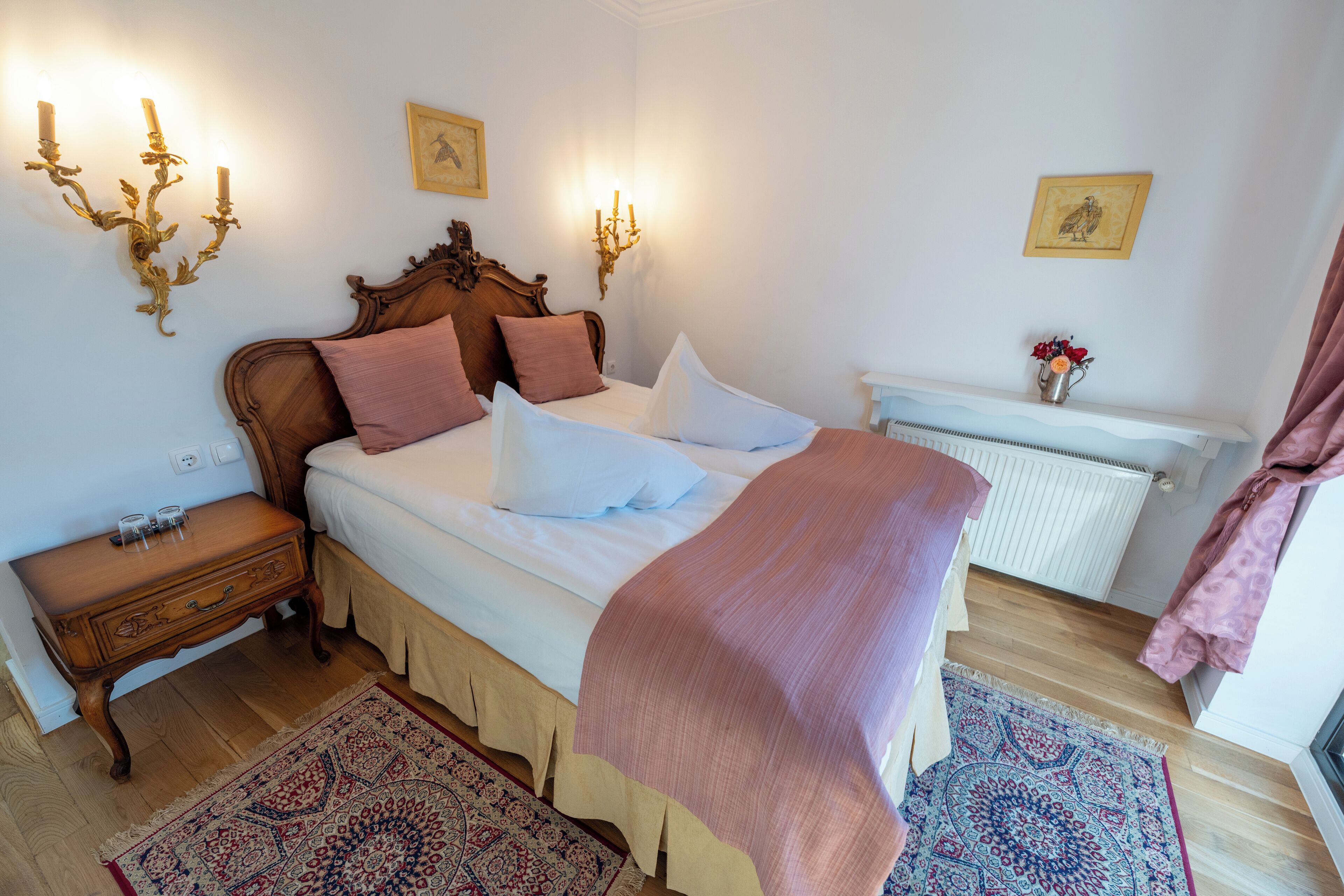 Une chambre à coucher avec un lit en bois, du linge de lit blanc, une table de chevet, une commode, un tapis à motifs et un radiateur.