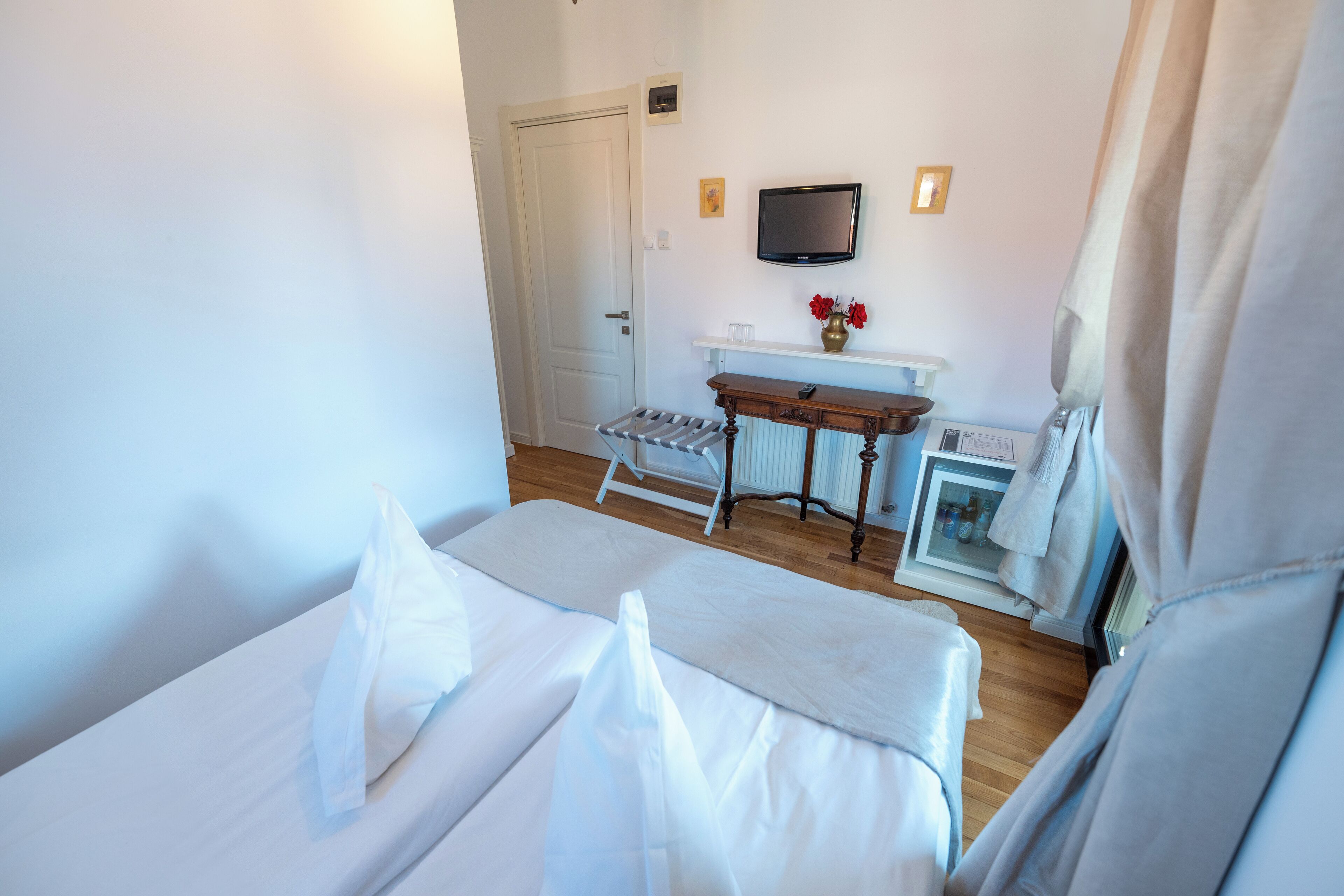 A hotel room with a bed, a small table with a vase of flowers, a television mounted on the wall, and a radiator.