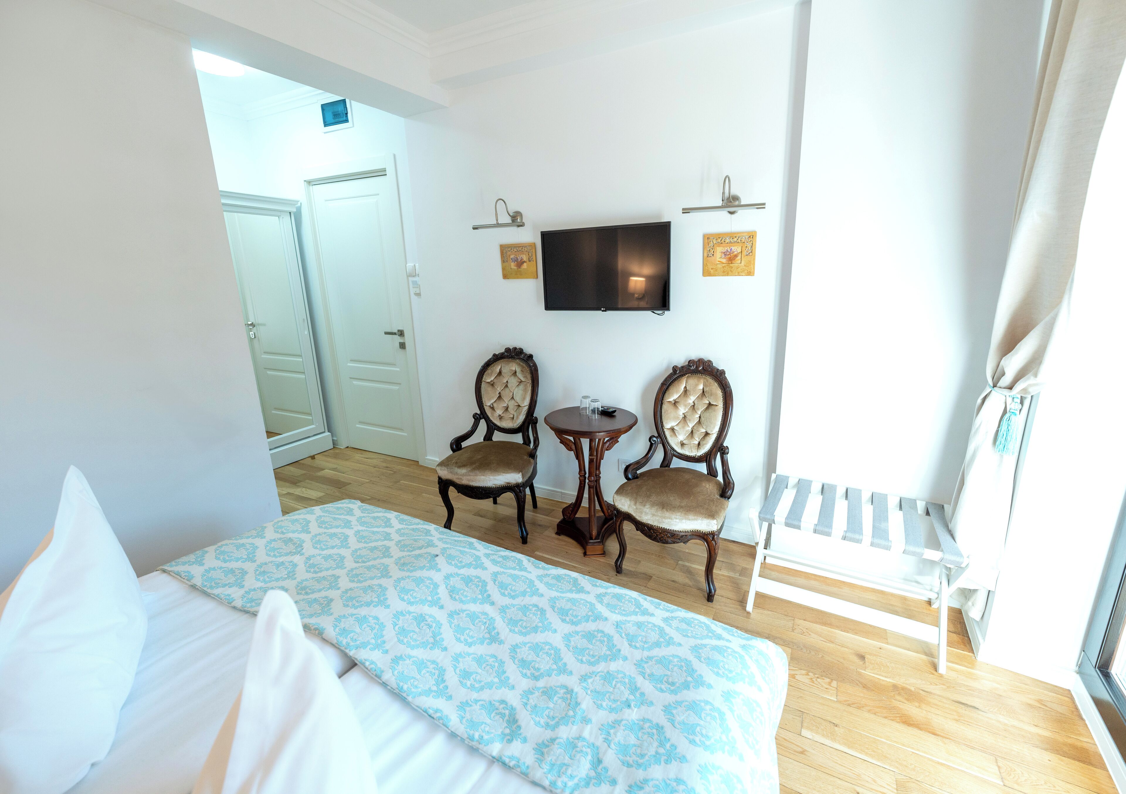 A hotel room with a bed, two chairs, a small table, a TV mounted on the wall, and a bench.