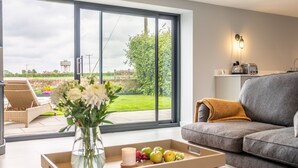 Un salon moderne avec un canapé gris, une table basse ornée d’un vase et de fruits, et une grande porte-fenêtre coulissante donnant sur un jardin.