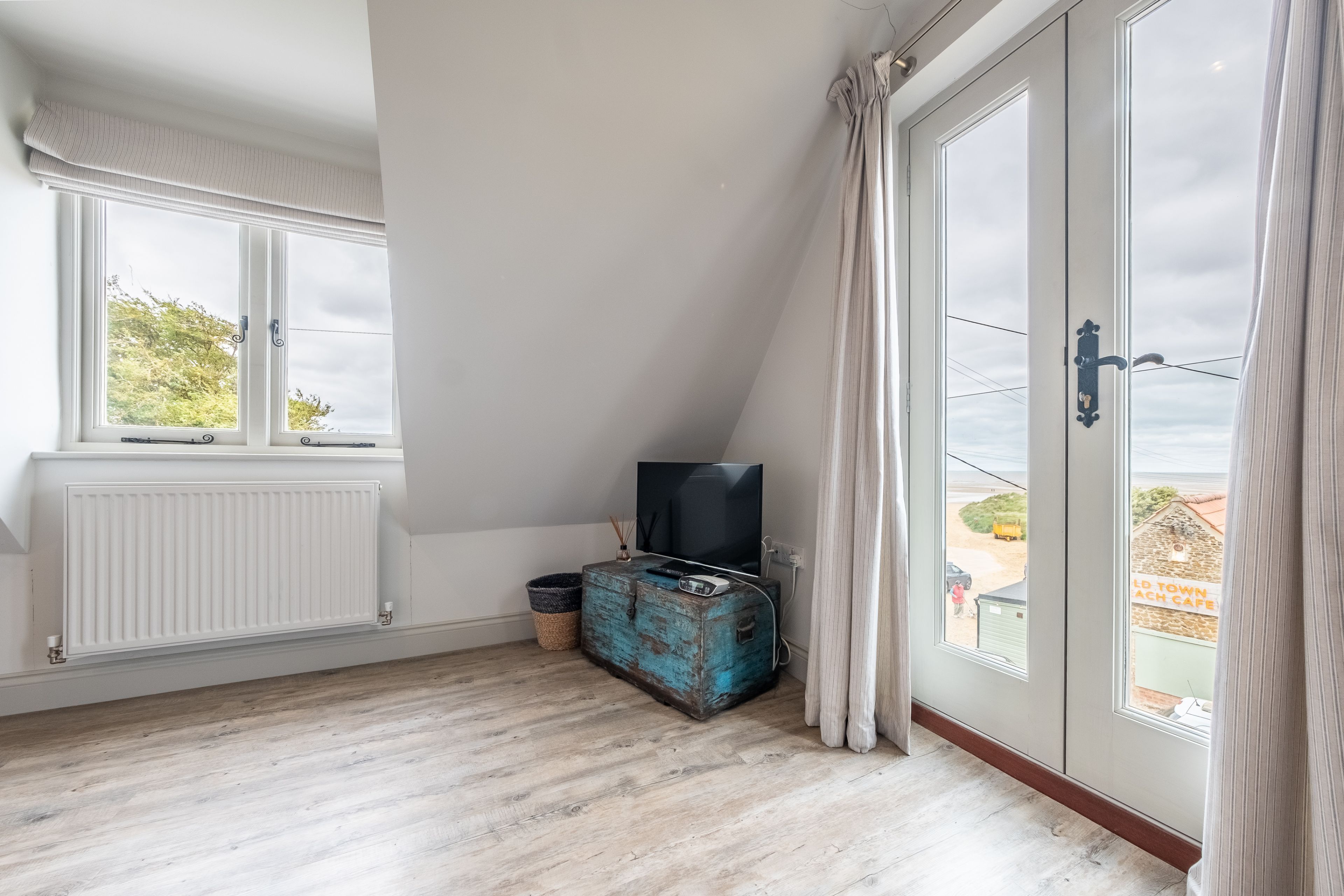 A room with a sloped ceiling, a white radiator, a television on a wooden chest, and a view of the outside through a glass door.