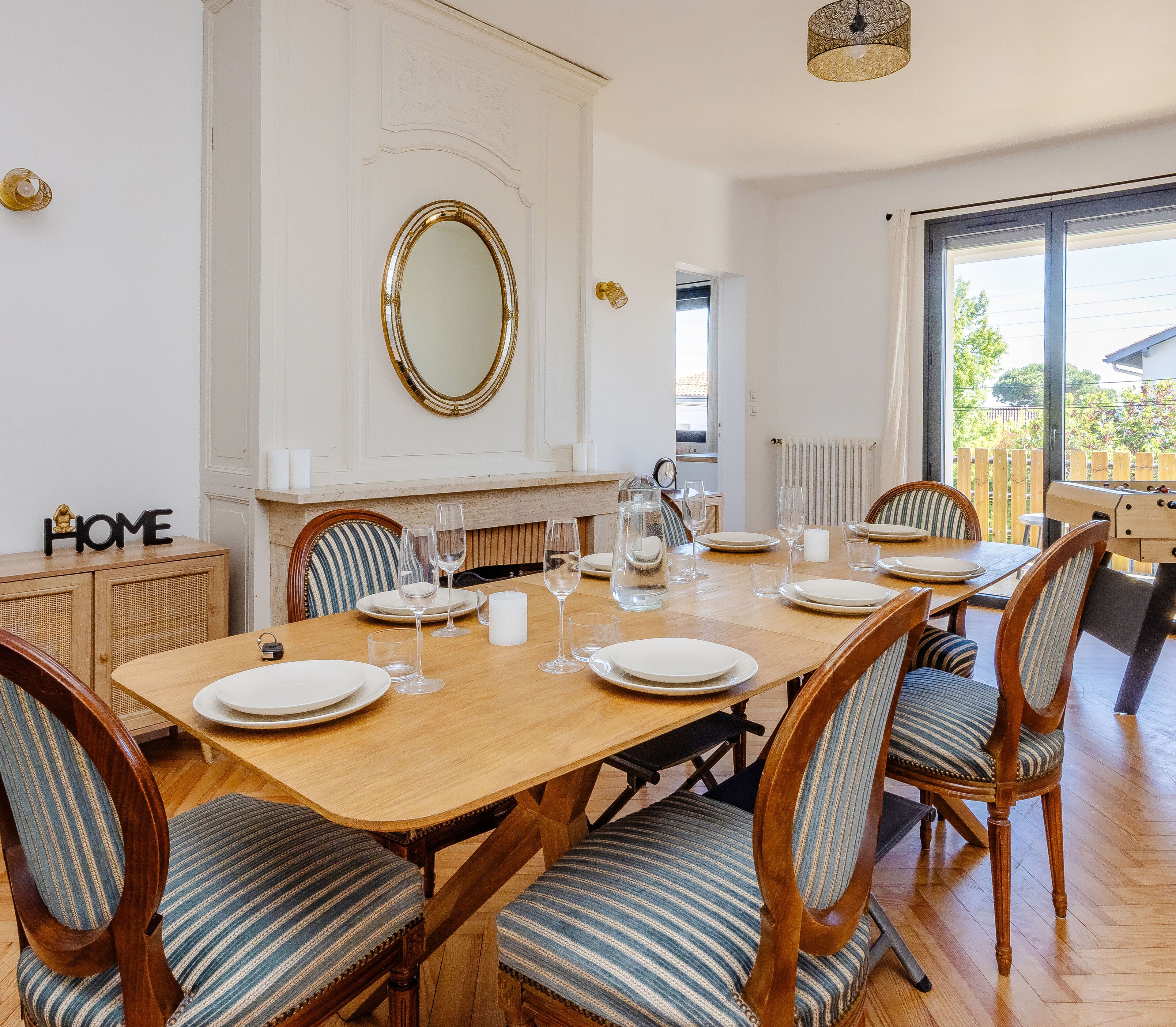 Un coin repas avec une table en bois dressée pour six personnes, entourée de chaises à motifs rayés. Un grand miroir est accroché au-dessus de la table, et on aperçoit une cheminée au fond.
