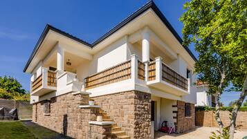 Une maison à deux étages avec une façade en pierre, un balcon doté de balustrades en bois et un ciel d’un bleu éclatant.
