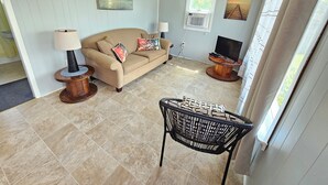 A living room with a beige sofa, a coffee table, a TV, and a window with blinds. - Lyons Cottages - Cottage #5, 2BR Walk to the Beaches of Stanhope National Park! (Stanhope)