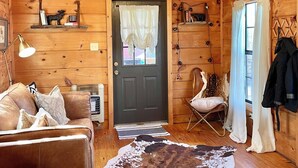 A cozy cabin interior with a wooden ceiling, a ceiling fan, a brown leather sofa, a cowhide rug, and a door with curtains. - Cozy cabin and contemporary container home on one property! Pets welcome! (Enoree)