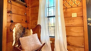 A cozy cabin interior with wooden walls, a wooden floor, a chair with a cushion, a small table with a decorative item, and a window with curtains. - Cozy cabin and contemporary container home on one property! Pets welcome! (Enoree)