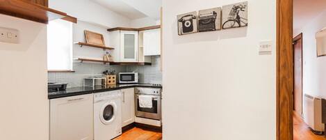 Une cuisine compacte avec des meubles blancs, un lave-linge, un four à micro-ondes et une cuisinière. On y trouve des étagères en bois au-dessus de l’évier et une œuvre d’art sur le thème du vélo accrochée au mur.