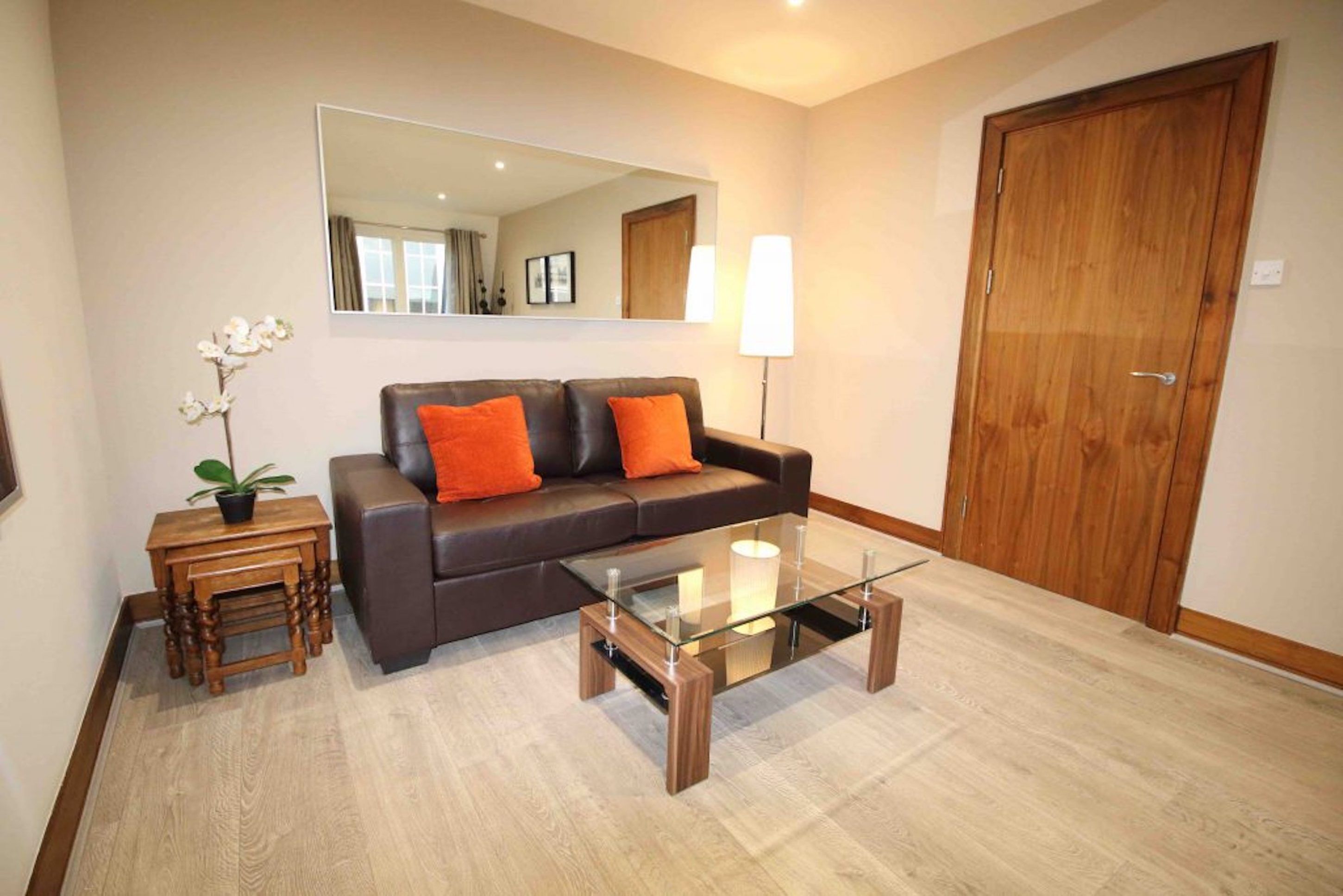 Een woonkamer met een donkerbruine leren bank, twee oranje kussens, een glazen salontafel, een houten bijzettafeltje met een plant, een vloerlamp en een houten deur.